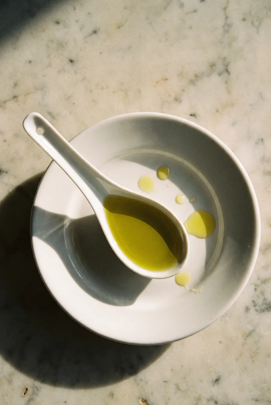 A tablespoon of golden green olive oil in a small white porcelain spoon on a white plate