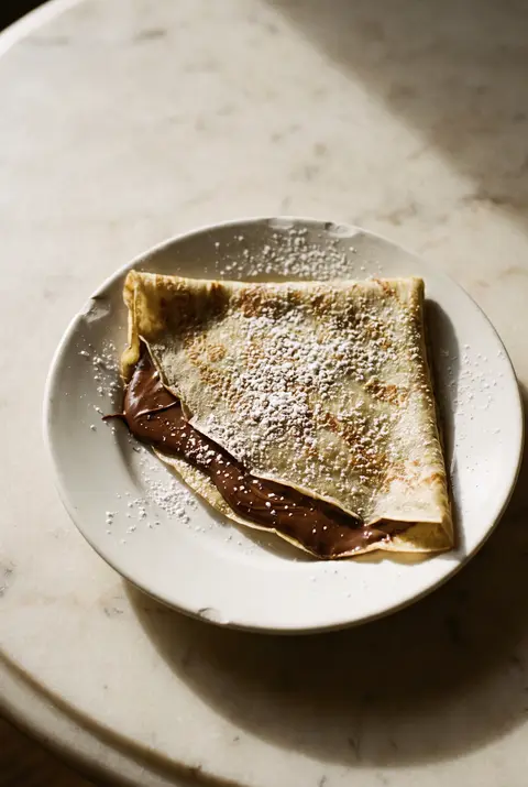 A thin crepe folded into quarters with chocolate hazelnut spread oozing out on a white plate