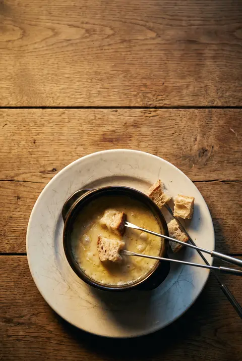 A small pot of melted cheese fondue with cubes of bread on long forks on a white plate