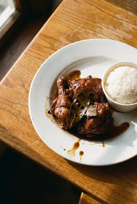 A plate of dark braised chicken adobo beside a cup of white rice on a white plate