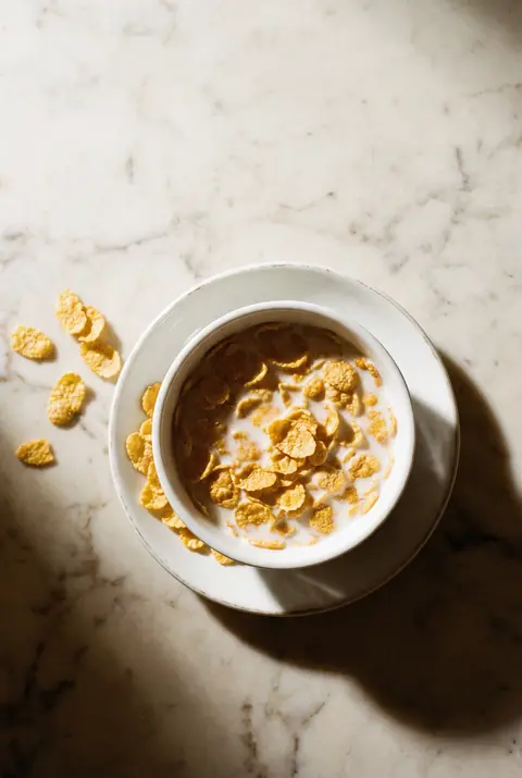A white porcelain bowl of cornflakes with milk on a white plate