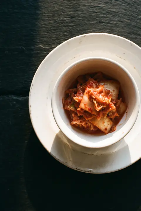 A side portion of red kimchi in a small white porcelain bowl on a white plate