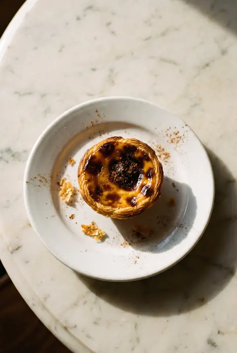 A Portuguese custard tart with a blistered dark caramel top and flaky pastry on a white plate