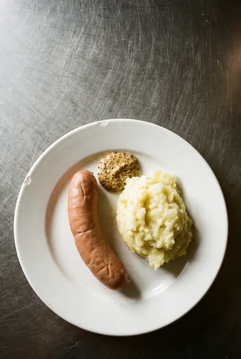 A boiled sausage with a scoop of mashed potato and a squeeze of mustard on a white plate