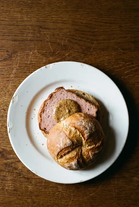 A thick slice of Bavarian Leberkase in a crisp bread roll with sweet mustard on a white plate