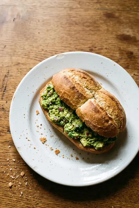 A Chilean marraqueta bread roll split open and filled with mashed avocado on a white plate