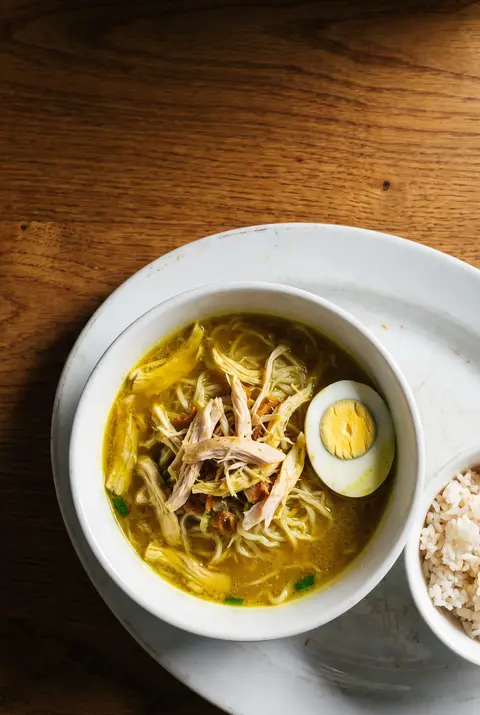 A white porcelain bowl of yellow soto ayam with shredded chicken on a white plate