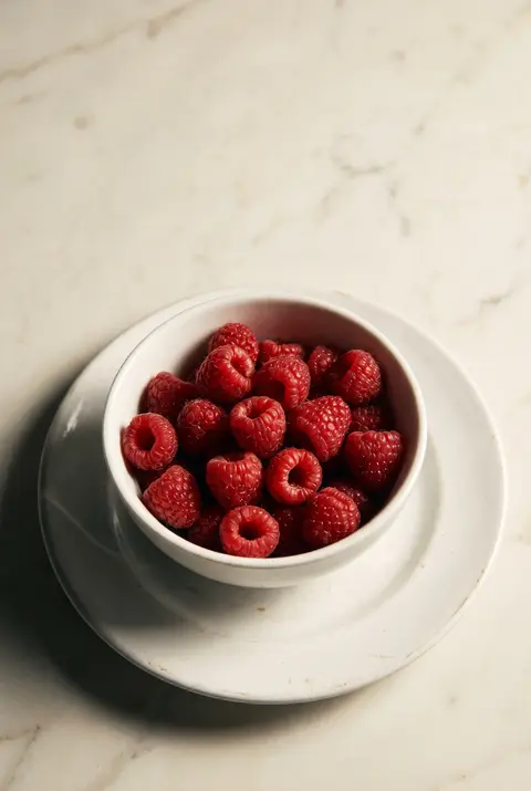 A white porcelain bowl of fresh raspberries on a white plate