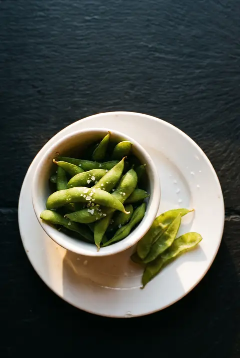 A small white porcelain bowl of edamame in their pods with flakes of sea salt on a white plate