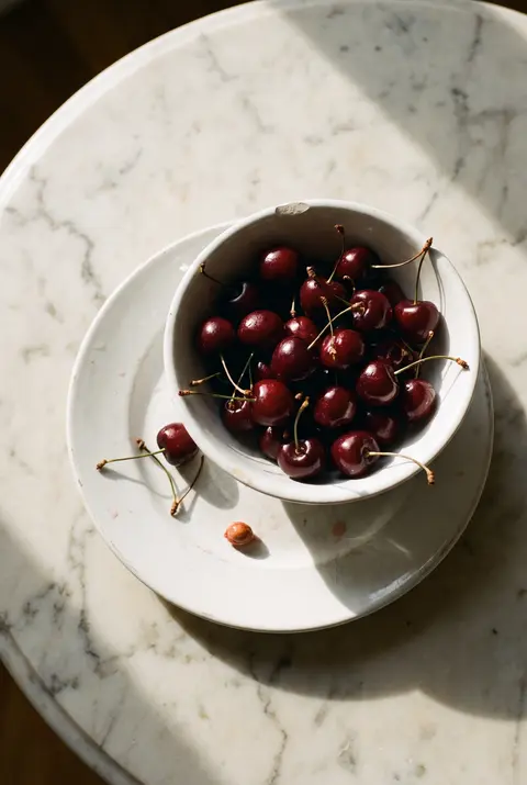 A white porcelain bowl of dark red cherries with stems on a white plate