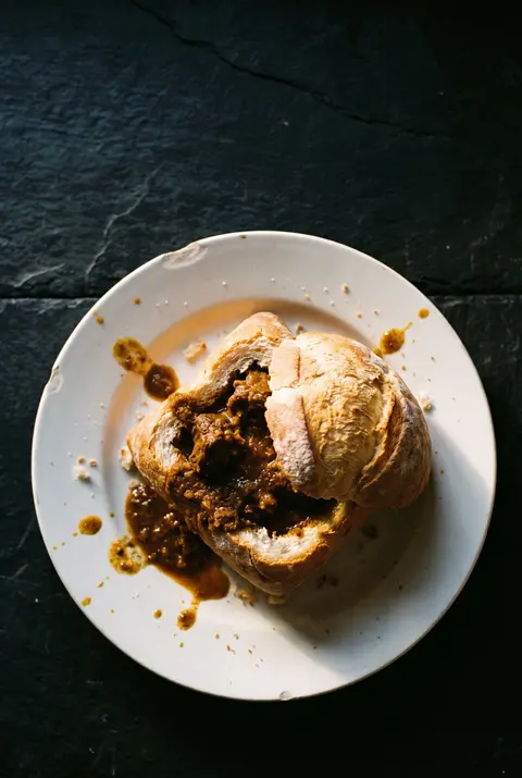A quarter loaf of white bread hollowed out and filled with mutton curry on a white plate