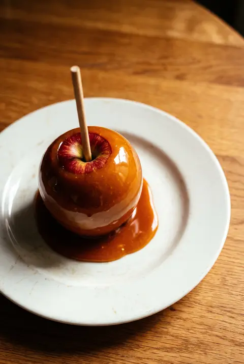 A apple coated in plain caramel on a wooden stick on a white plate