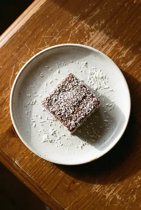 A square lamington coated in chocolate and coconut on a white plate