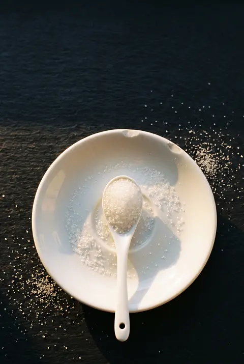 A teaspoon of white sugar on a small white porcelain spoon on a white plate