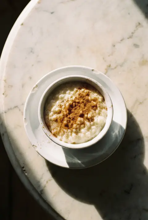A white porcelain bowl of creamy rice pudding with a dusting of cinnamon on a white plate