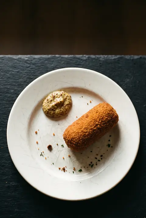 A golden Dutch kroket with a spoonful of mustard on a white plate