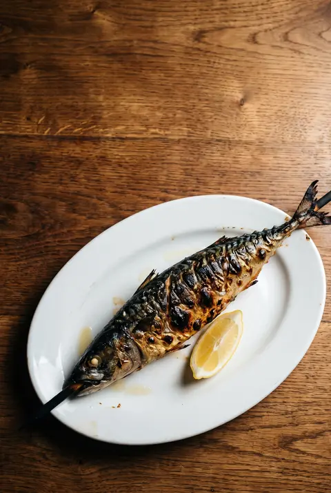 A whole grilled mackerel on a wooden stick with charred skin on a white plate