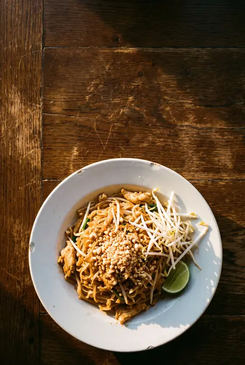 A takeaway size plate of chicken pad thai with crushed peanuts and a wedge of lime on a white plate