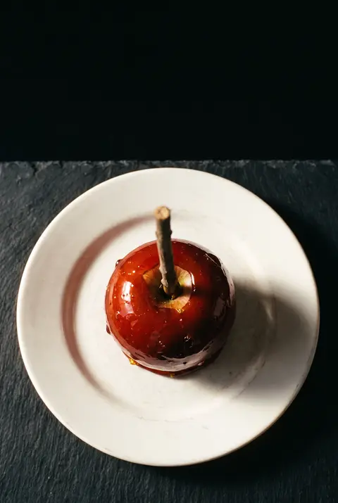 A red toffee apple with a hard red caramel shell on a wooden stick on a white plate