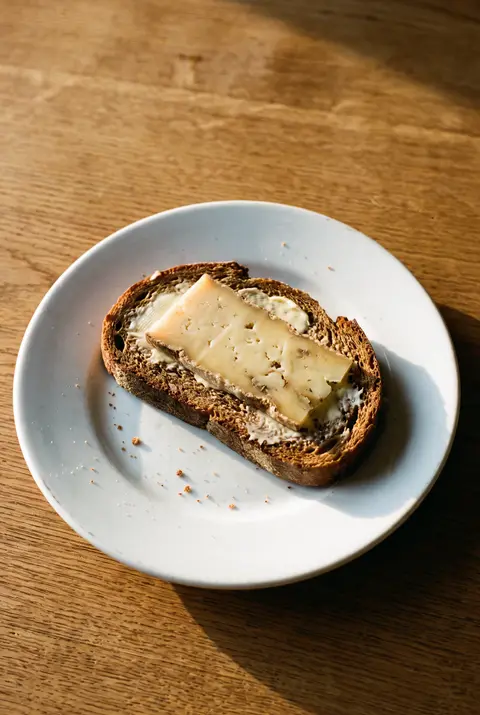 A slice of dark rye bread with butter and a slice of cheese on a white plate