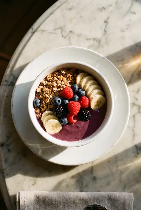 A white porcelain bowl of purple acai topped with granola on a white plate